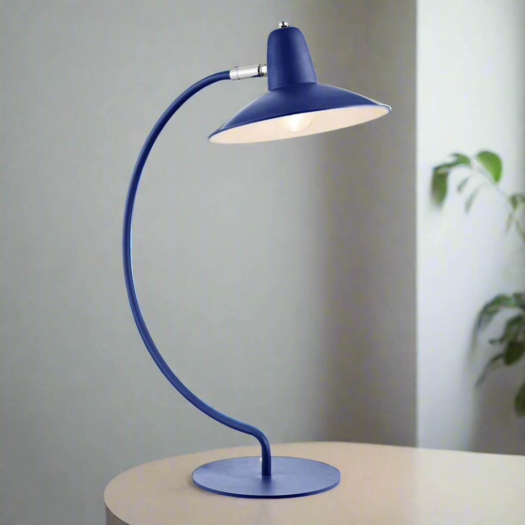 An arc shaped metal table lamp in a matte blue painted finish. The lamp is shown with a bulb just visible and is turned on in this picture. The lamp is placed on a side table with a green plant in the background.