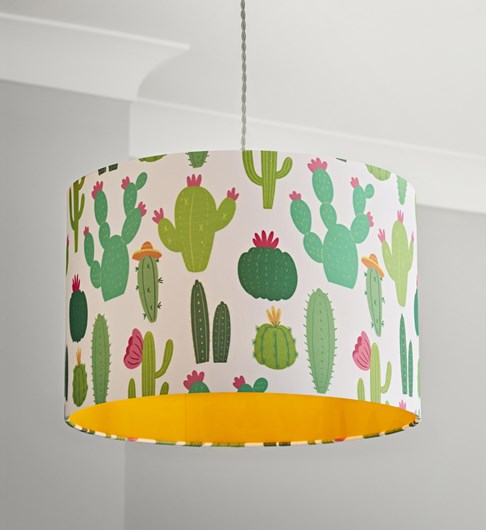 A picture of a straight sided white cotton cylinder lampshade that is covered in printed cartoon style illustrations of various different types of cactus plants. Some are dark green with ink flowers, some are pale green with orange flowers. Once of the cactus plants is wearing an orange coloured sombrero style hat. The lampshade has an orange coloured fabric lining. The picture is taken in a room with pale grey walls and white cornice.