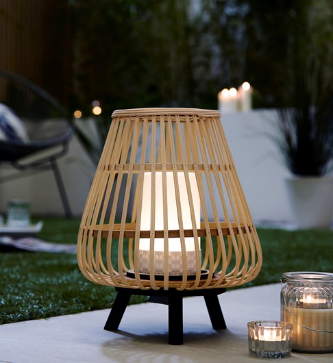 An outdoor shot if a bamboo cage style lantern style table lamp stood on a black coloured tripod base. The lantern has a white plastic pillar candle style central piece which houses a battery powered LED bulb that is turned on in this picture. The picture is taken at dusk on a garden patio with 2 small tealight candles to the right and an out of focus garden armchair in the backgroud to the left.