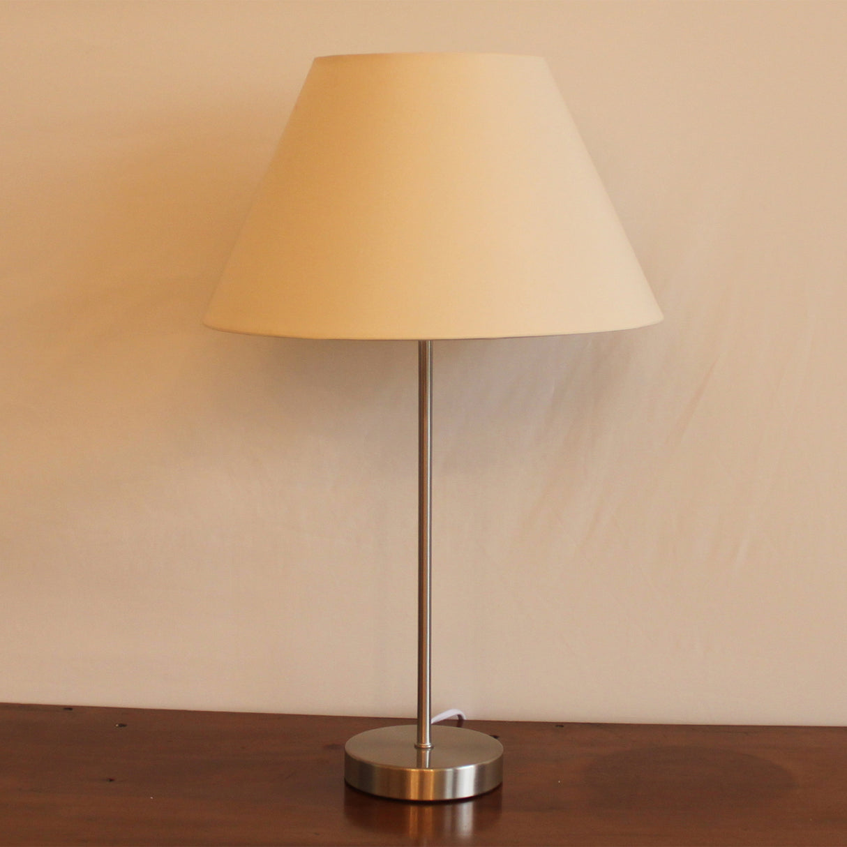 A picture of a chrome metal table lamp base with a tapered cream coloured cotton lampshade stood on top of a wooden furniture unit. 