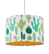 A picture of a straight sided white cotton cylinder lampshade that is covered in printed cartoon style illustrations of various different types of cactus plants. Some are dark green with ink flowers, some are pale green with orange flowers. Once of the cactus plants is wearing an orange coloured sombrero style hat. The lampshade has an orange coloured fabric lining. The picture is taken against a white background.
