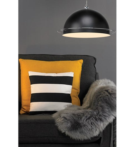 A retro looking black lampshade hanging on a grey coloured twisted ceiling cable. The dome shaped lampshade has a shiny chrome ring approximately 2.5cm or 1 inch from the bottom. The lampshade is shown hanging low above a dark coloured fabric armchair with a fur throw over one side and a large yellow and small black and white cushion.